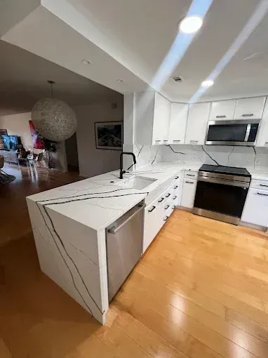 A modern white kitchen with marble-veined quartz countertops, stainless steel appliances, and light wood flooring.