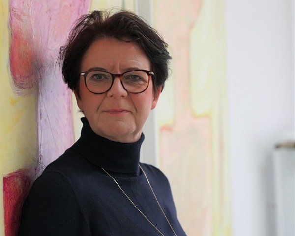 Frau mit Brille und dunklem Rollkragenpullover, lehnt an einem farbenfrohen abstrakten Gemälde.