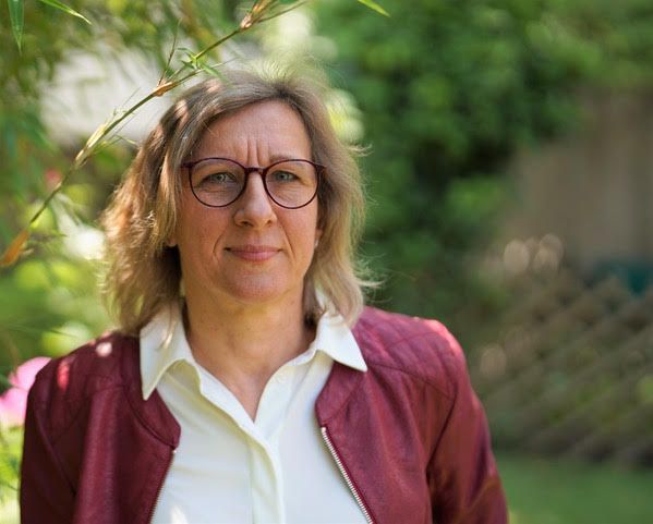 Frau mit Brille, roter Jacke, weißem Hemd, in einem Garten.