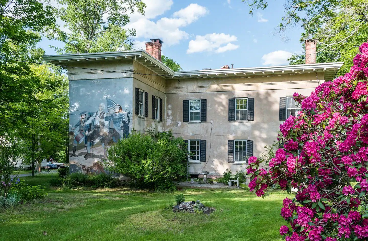 A large house with a mural on the side of it is surrounded by trees and flowers.