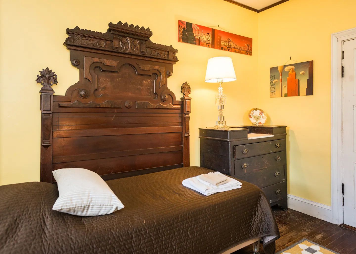 A bedroom with a single bed , dresser , lamp and paintings on the wall.