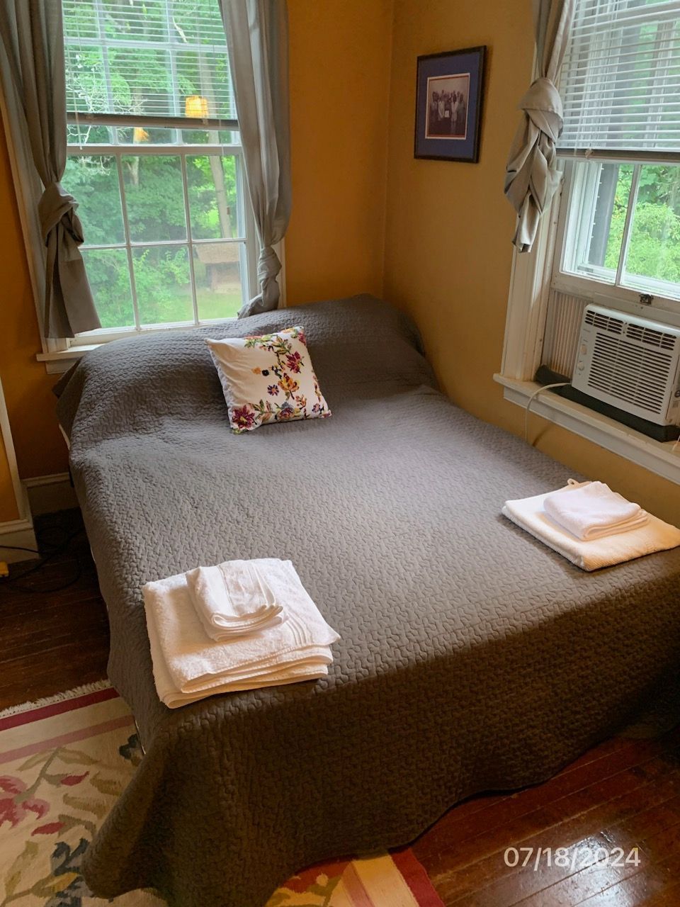 Bedroom with a bed, towels, two windows with gray curtains, and an air conditioner.