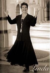 A woman in a black dress is standing in front of a building with her arms outstretched.