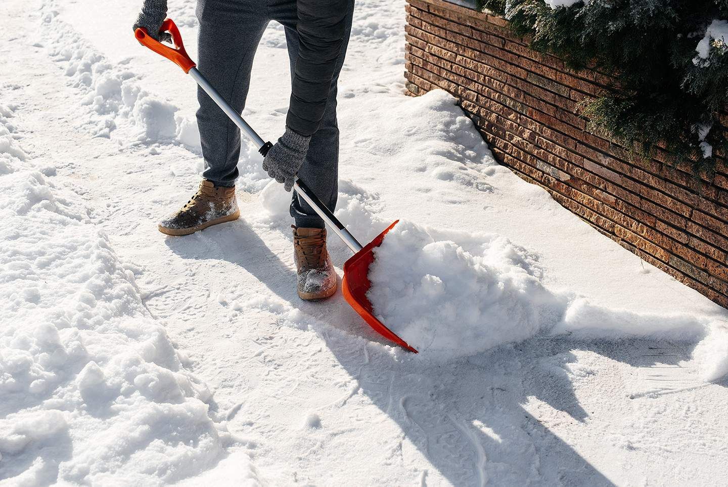 Person shoveling snow on a snowy sidewalk, using an orange shovel, near a wooden fence.