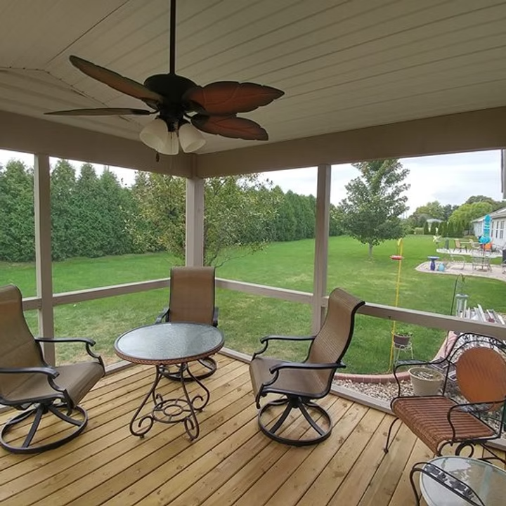 Covered porch with rocking chairs, round table, and ceiling fan overlooking a green yard and pool.