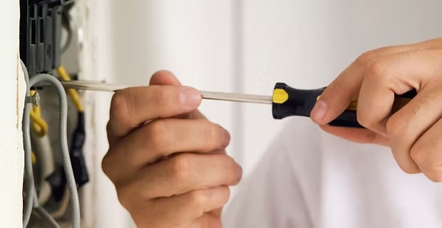 Hands using a screwdriver to tighten a wall fixture near hanging tools.