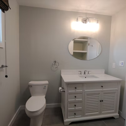 Small bathroom with toilet, vanity sink, round mirror, and wall light in light gray decor
