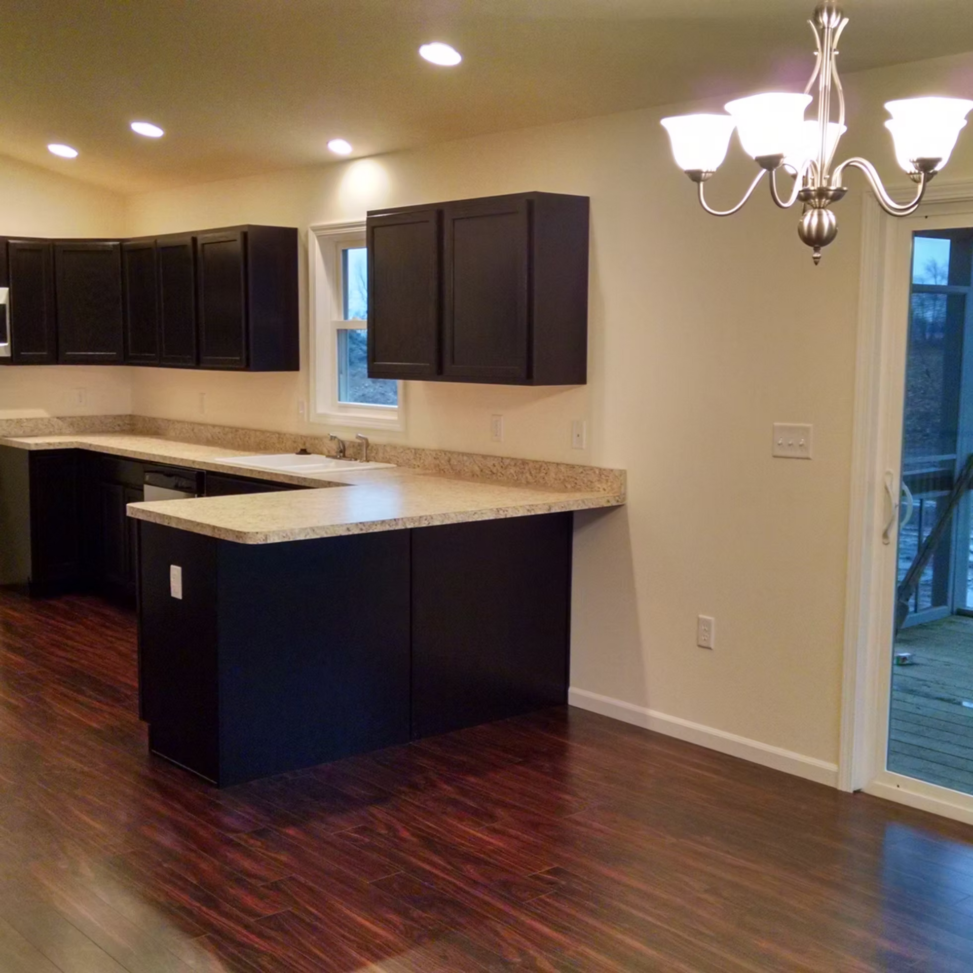 Empty modern kitchen with dark cabinets, light countertops, hardwood floors, and a chandelier