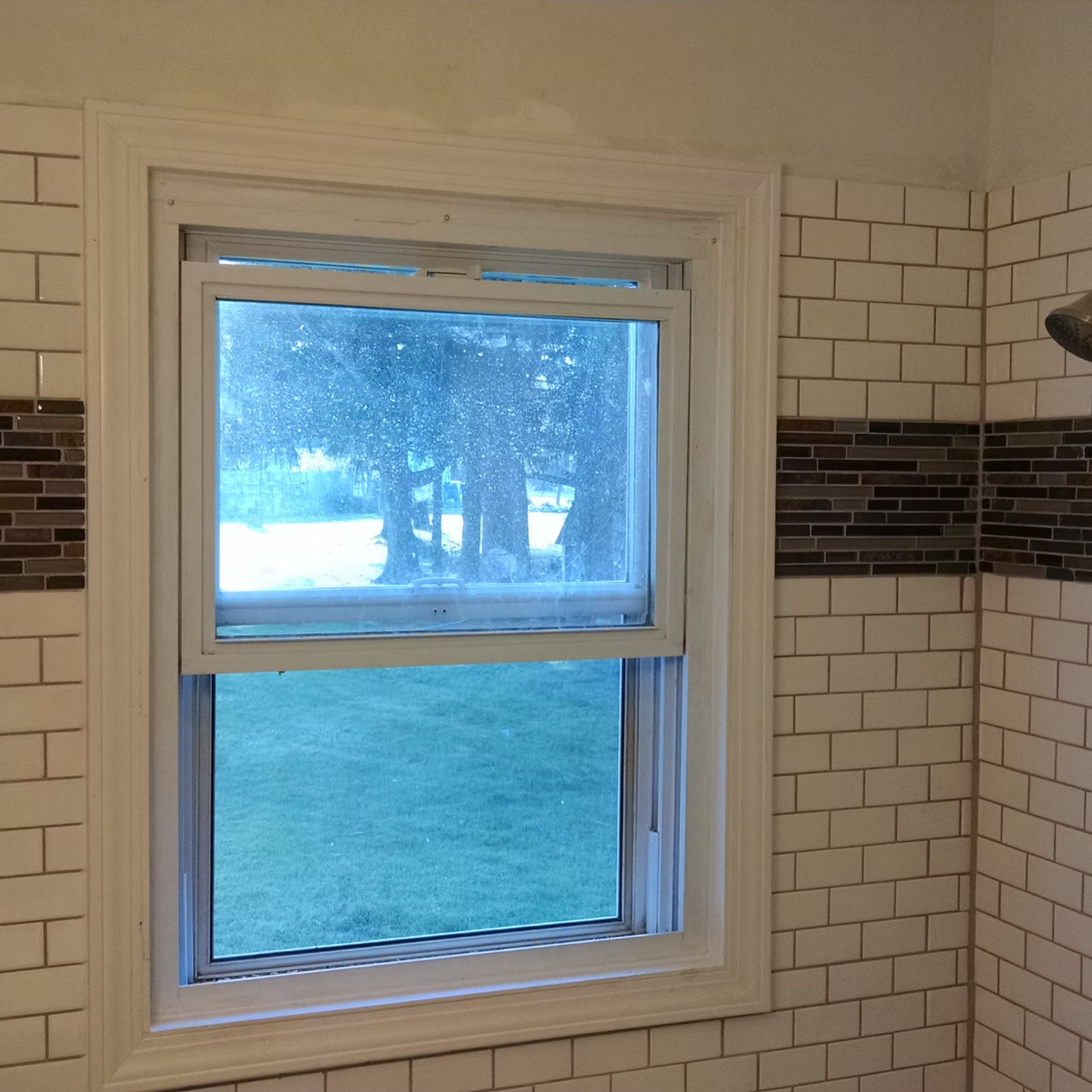 Bathroom window above a shower tile wall, showing a grassy yard and trees outside.