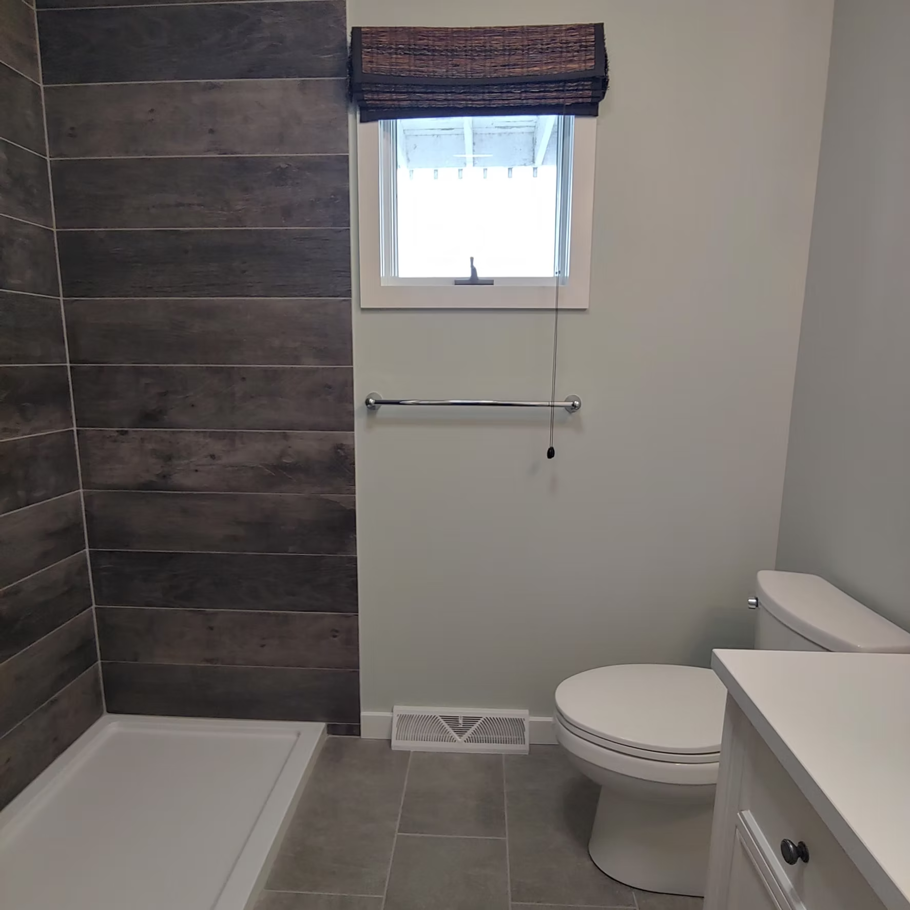 Small bathroom with shower, toilet, window, towel bar, and gray tile walls and floor.