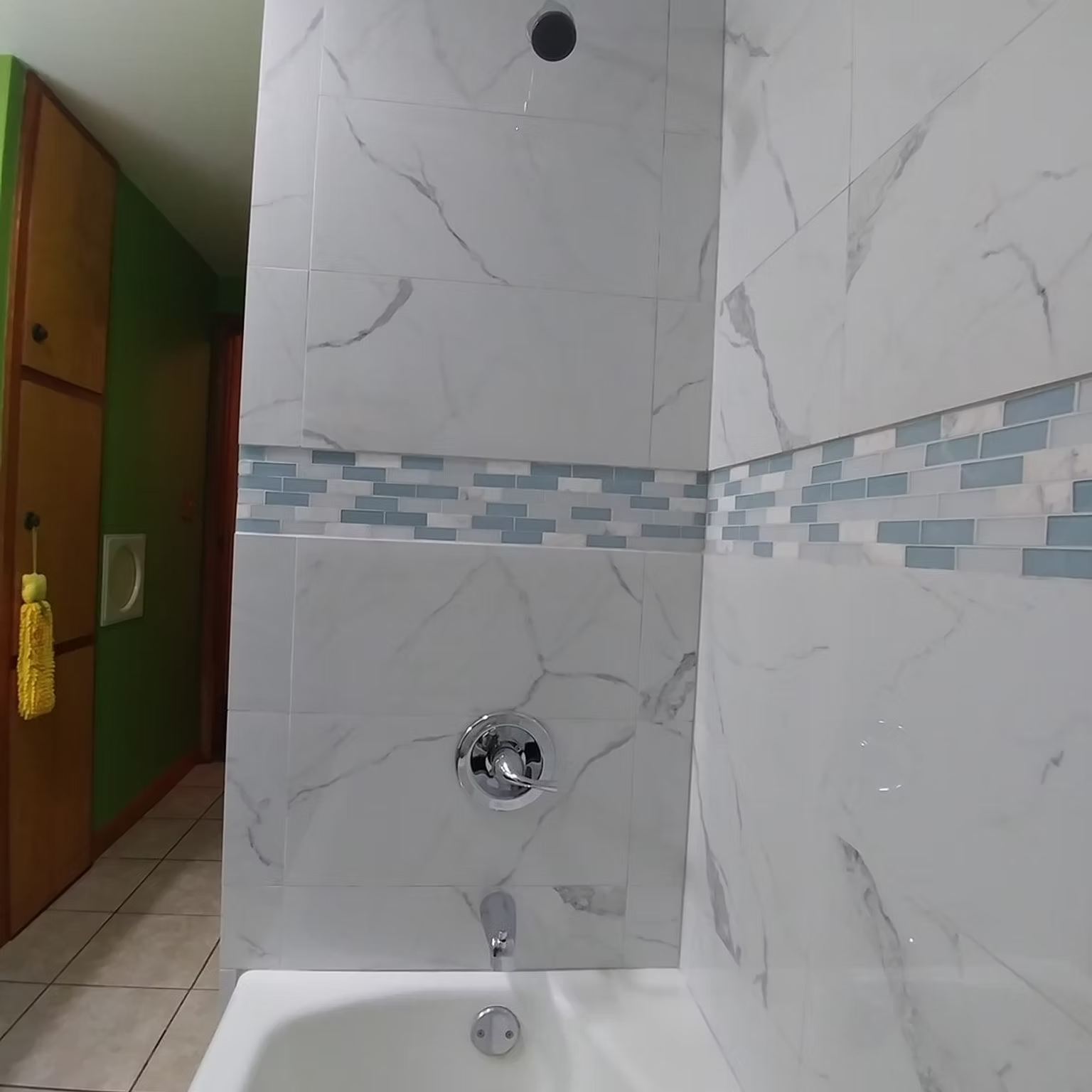 White tiled bathroom shower with blue mosaic stripe, chrome fixtures, and wooden door at left