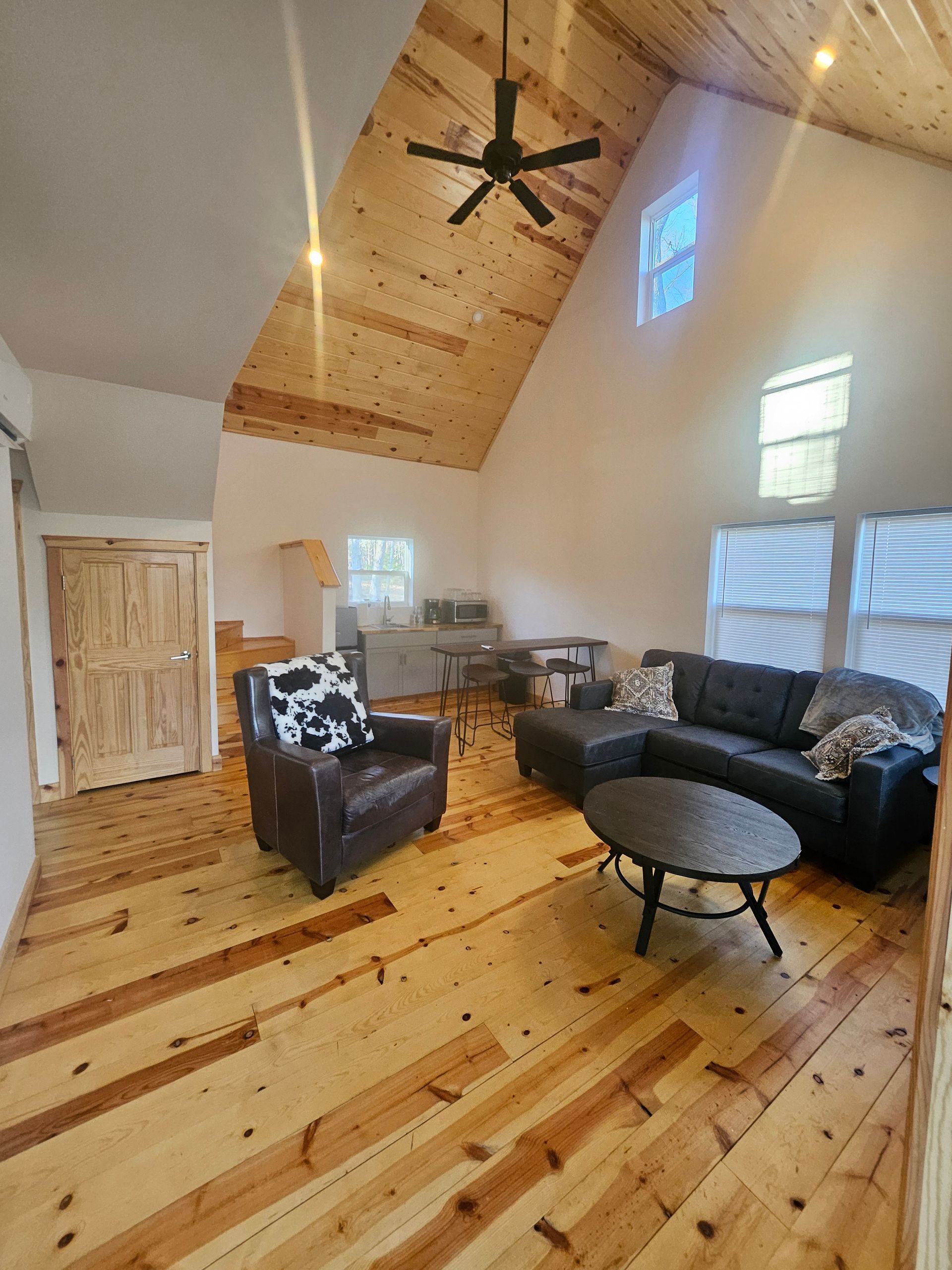 Bright living room with wood floors, black sofa and chair, round coffee table, and vaulted ceiling with fan