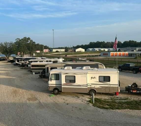 Several RVs parked along a gravel lot near a road, with a field and American flag in the distance.