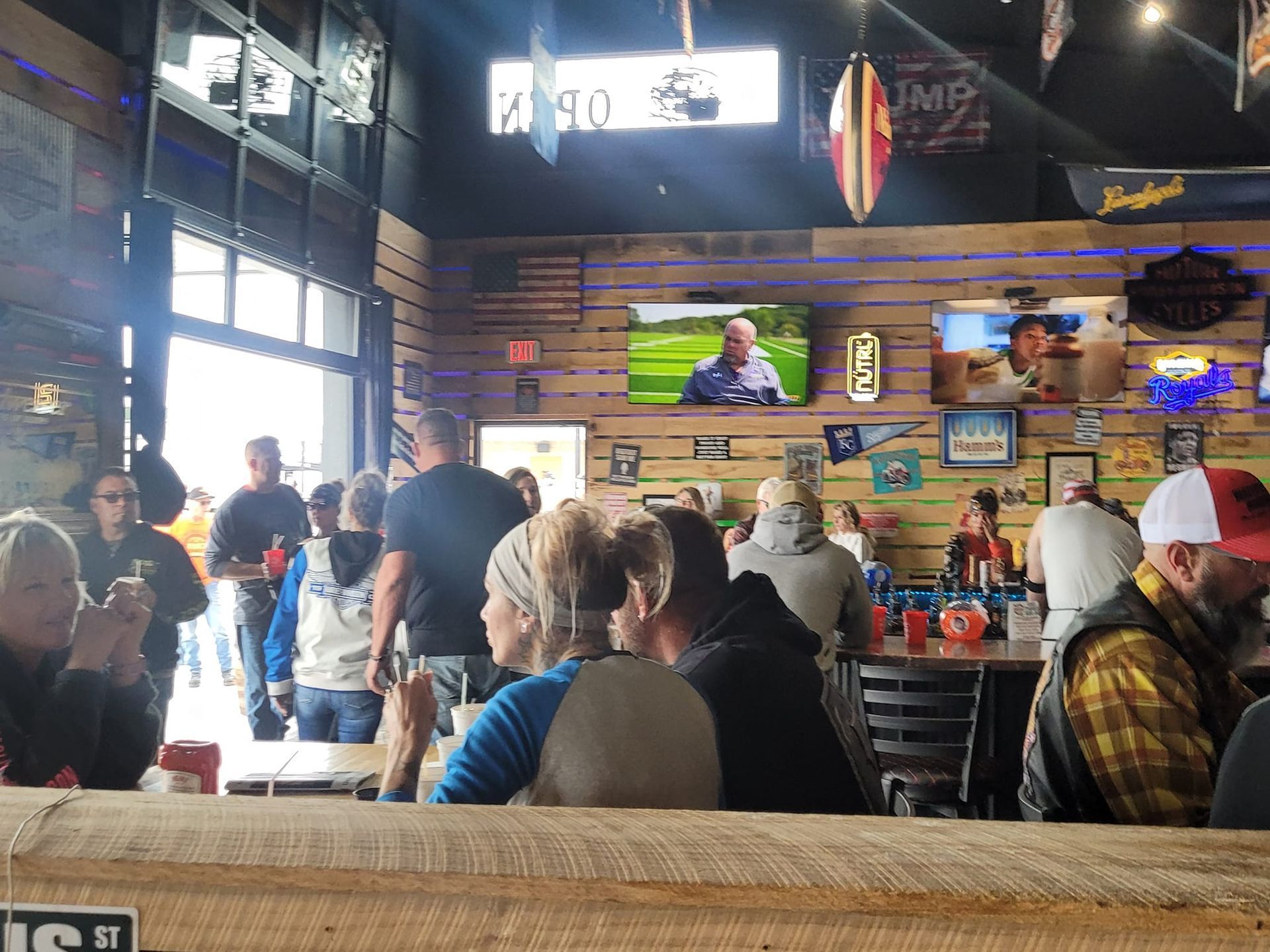 Busy rustic restaurant with patrons seated at tables and a server TV above the bar.