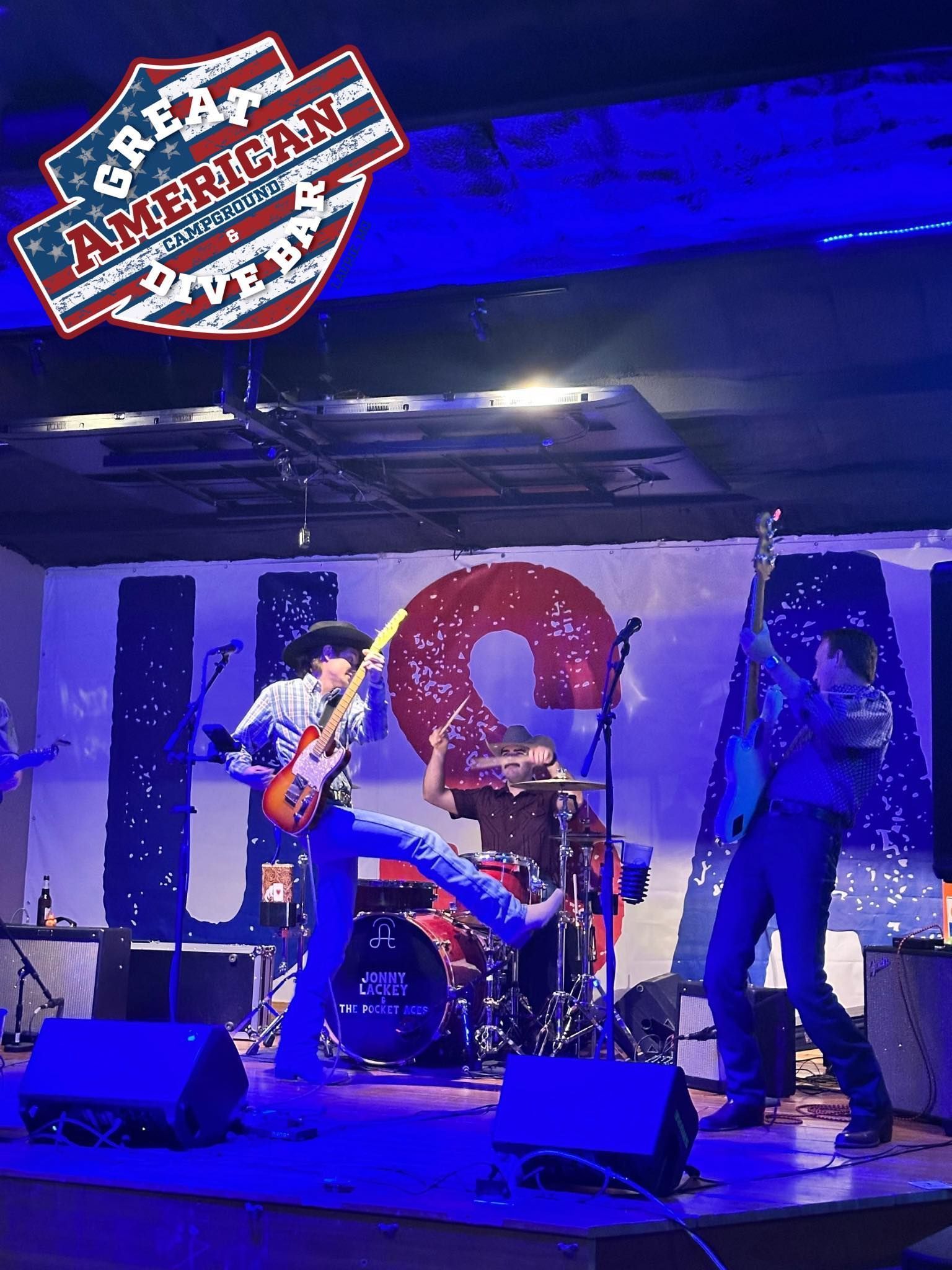 Band performing on a blue-lit stage at an American-themed bar, with drums, guitars, and large mural backdrop.