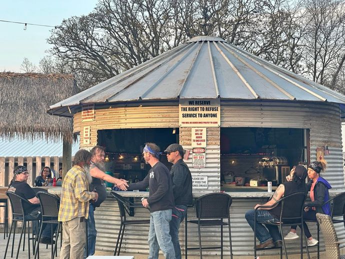 People chatting at an outdoor bar kiosk with a metal roof and stools by the water