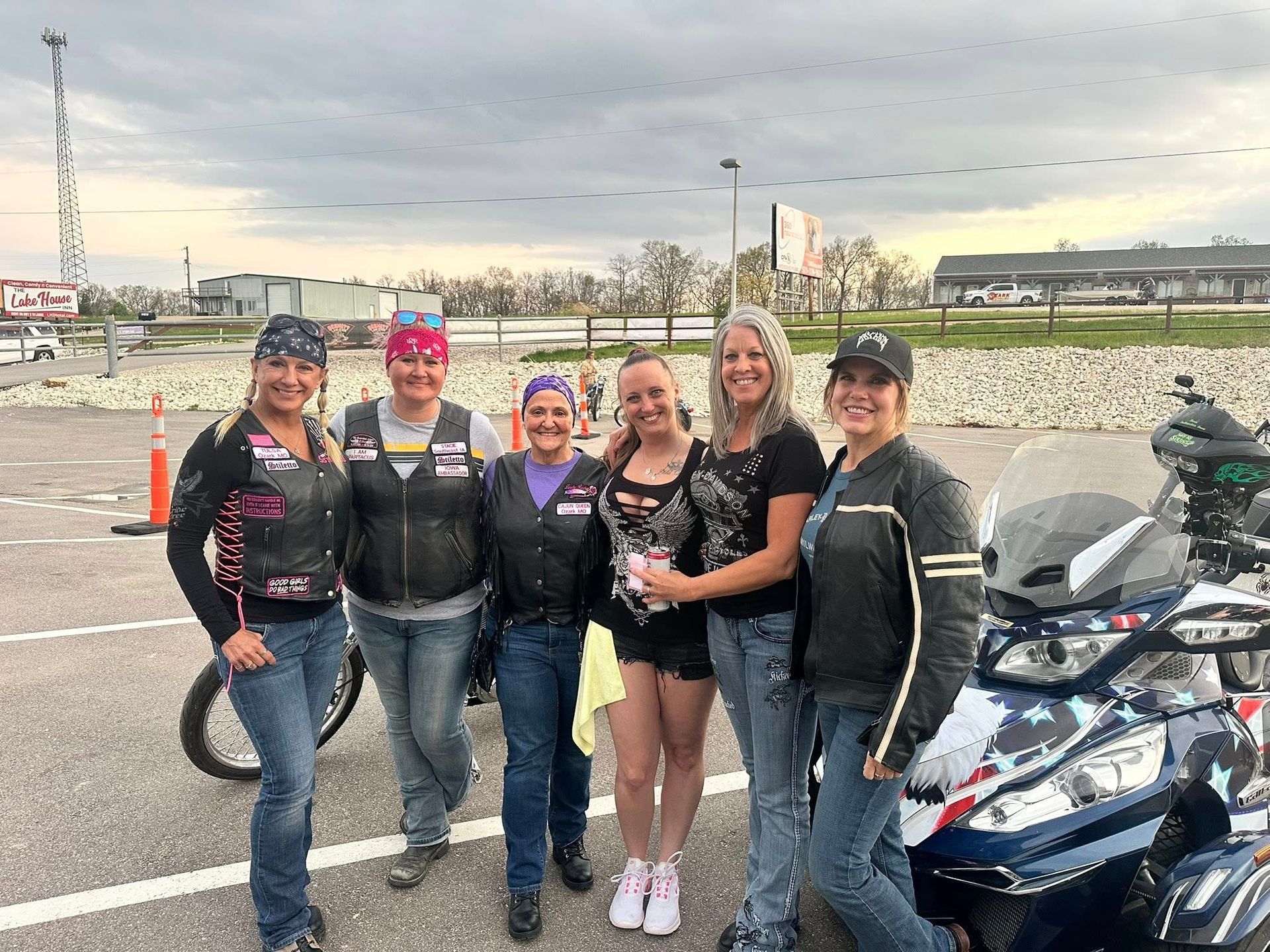 Five women posing outdoors beside a parked motorcycle on a cloudy day.