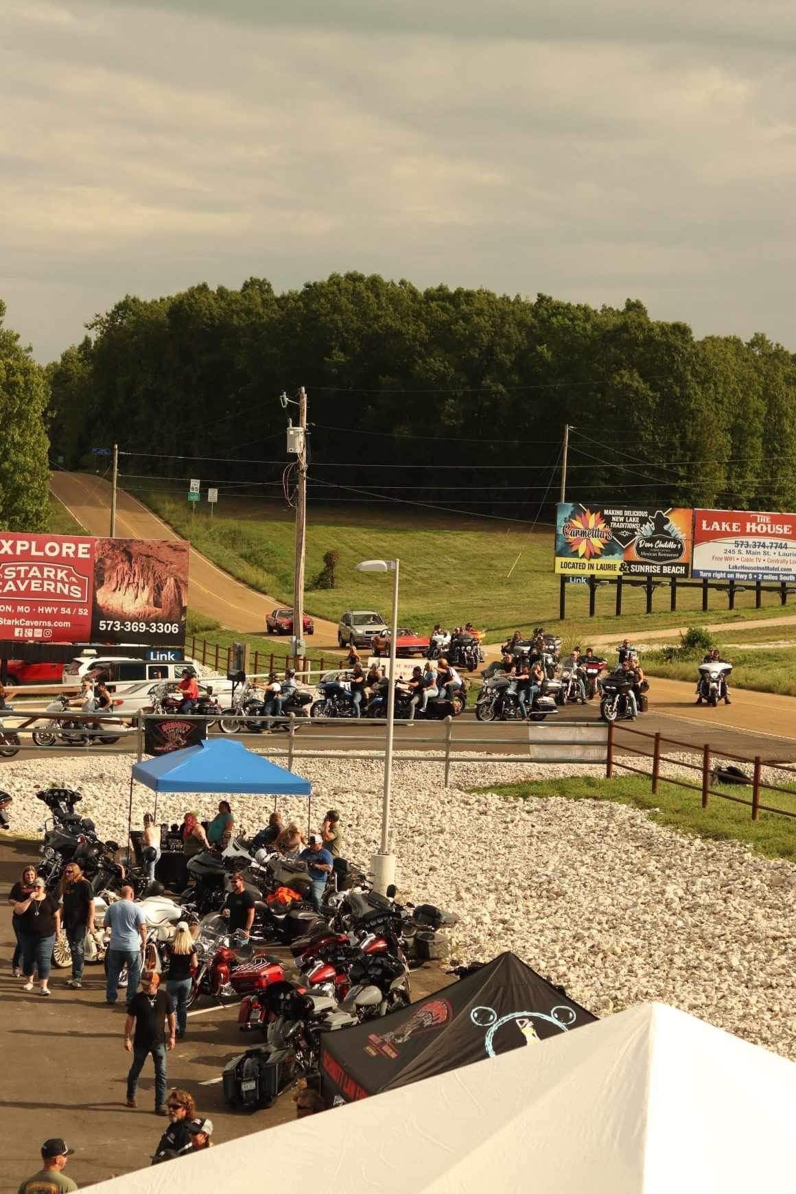 Motorcycles lined up at an outdoor rally near a dirt track and green hillside under cloudy skies