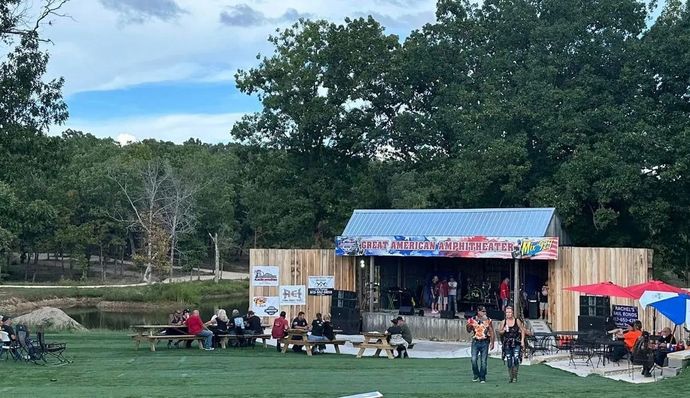 Outdoor stage at a park festival with performers, seated crowd, trees, and red umbrellas by a pond