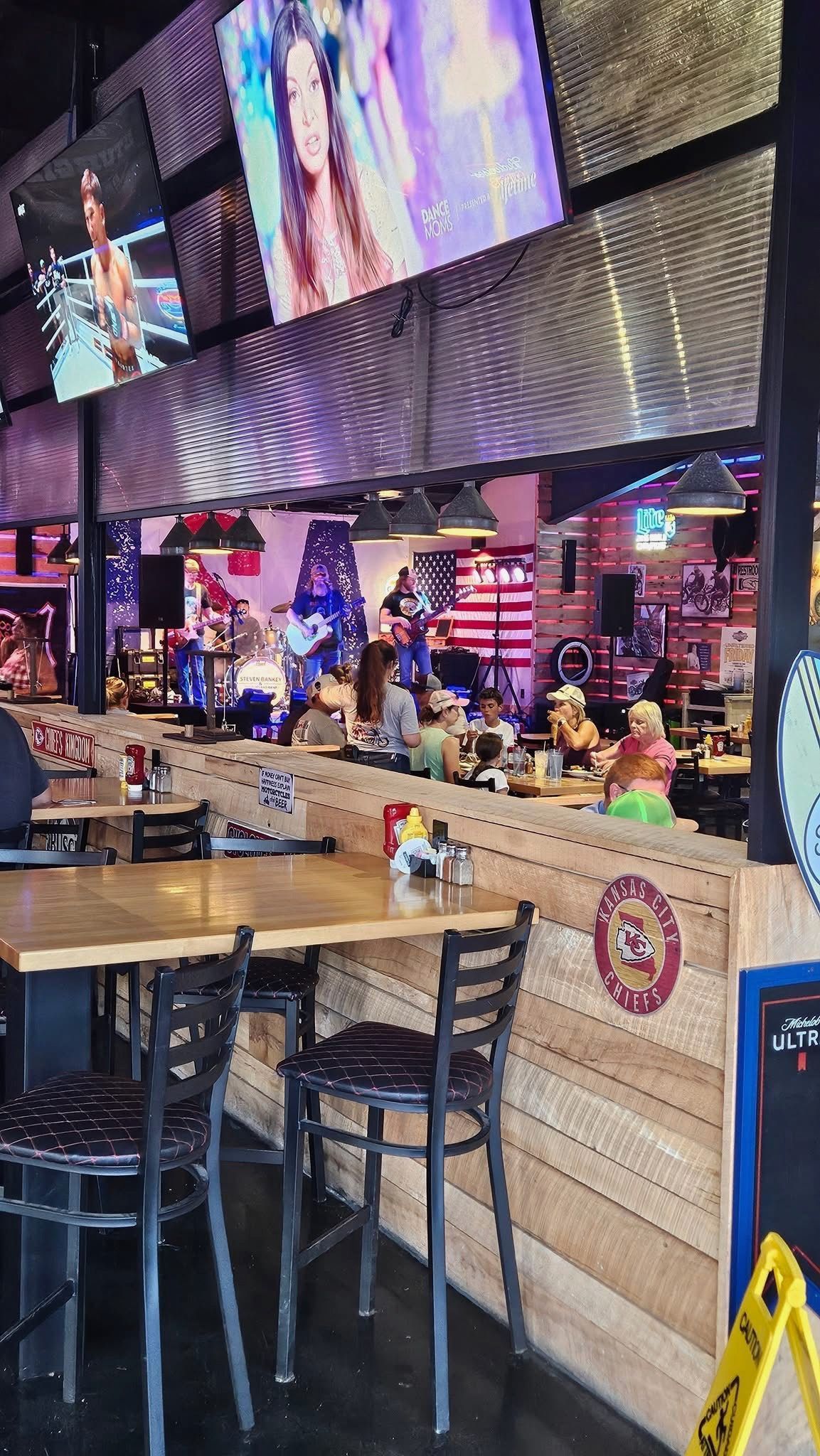 Sports bar with a long wooden table, bar stools, large screens, and neon-lit TV walls.