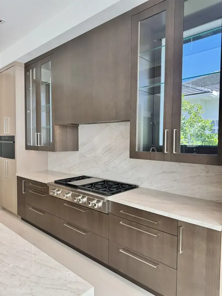 Dark oak kitchen cabinets with glass display uppers, full-height quartzite backsplash, and professional gas rangetop by Kitchen & Bath Studios.