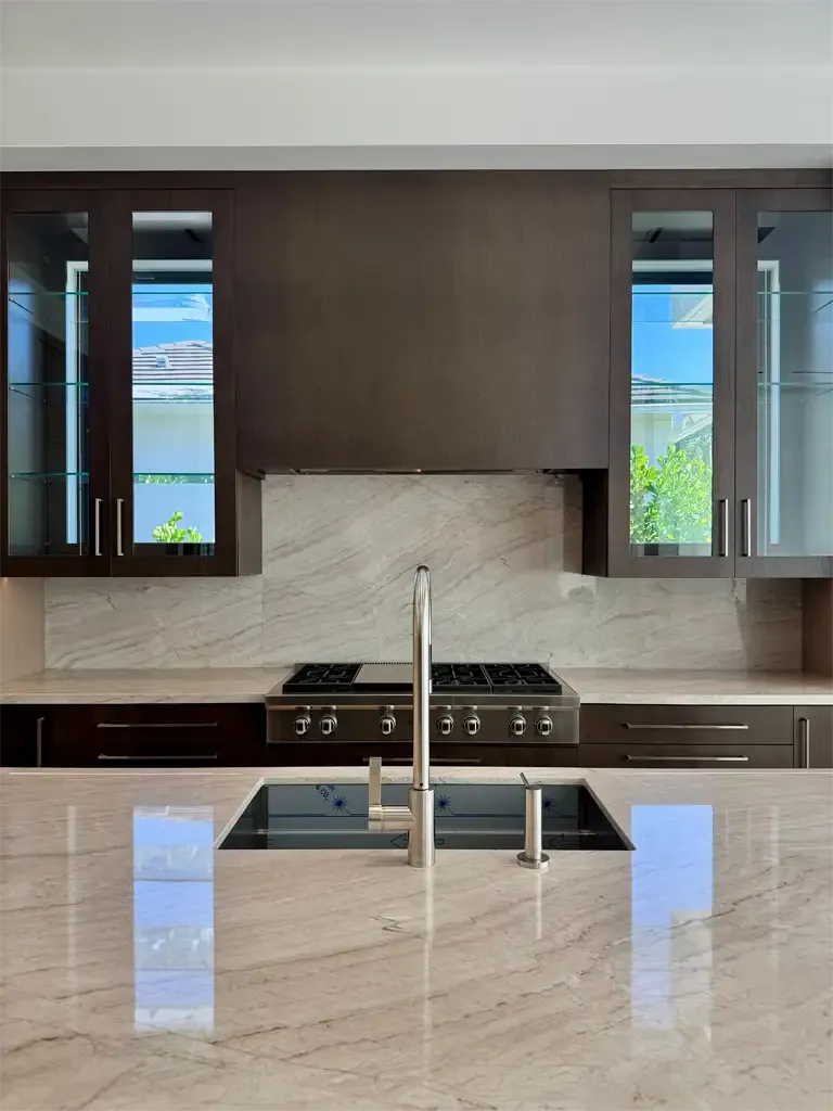 Custom espresso kitchen cabinetry with quartzite backsplash, glass-front cabinets, and island prep sink in South Florida home.