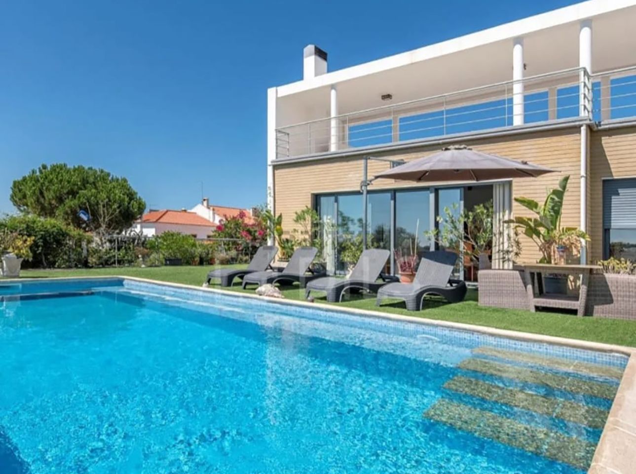 Casa moderna com piscina no quintal, espreguiçadeiras e água azul cristalina sob um céu ensolarado.