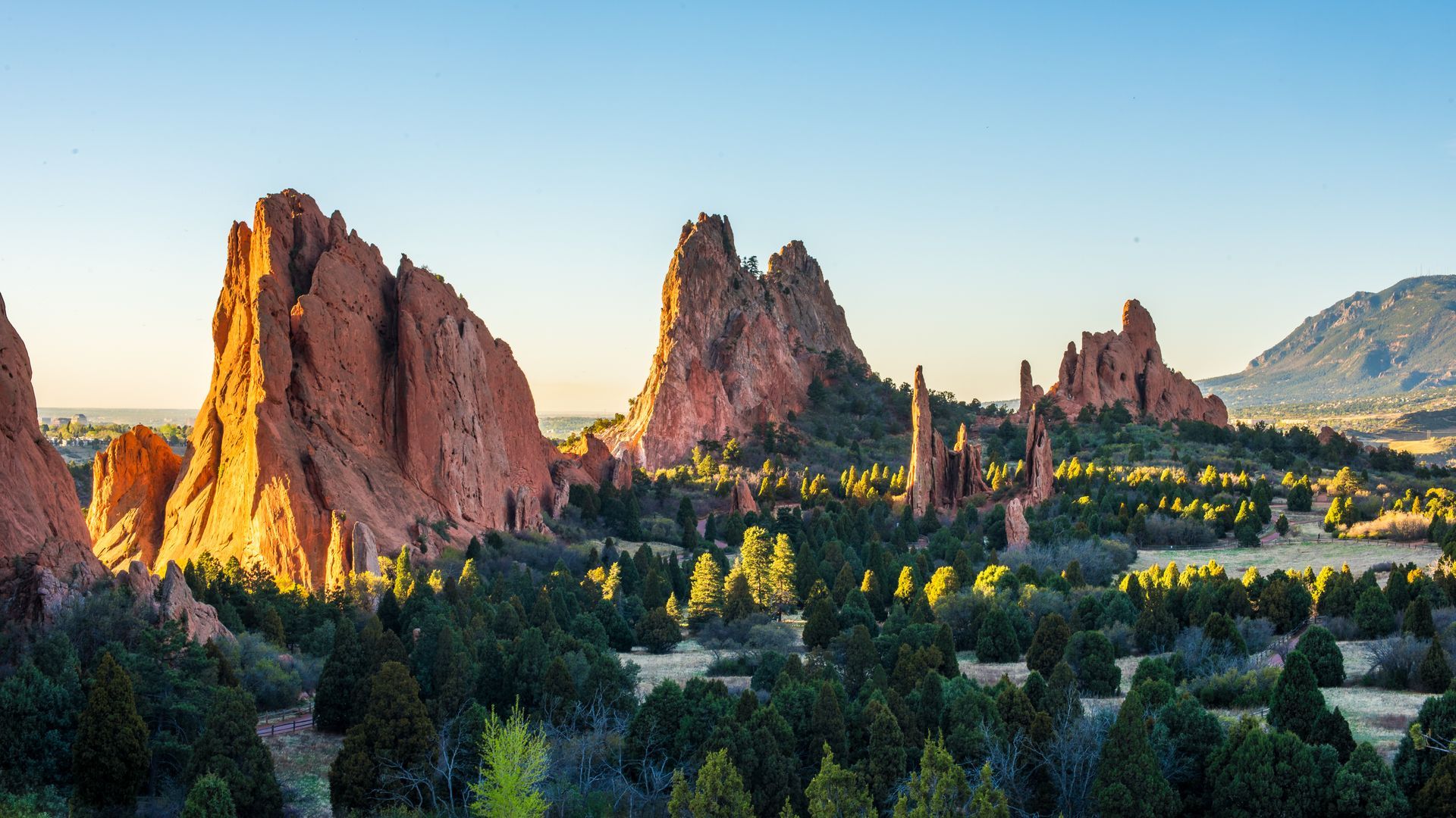 Sunlit red rock spires above a green valley at sunrise, with mountains in the distance