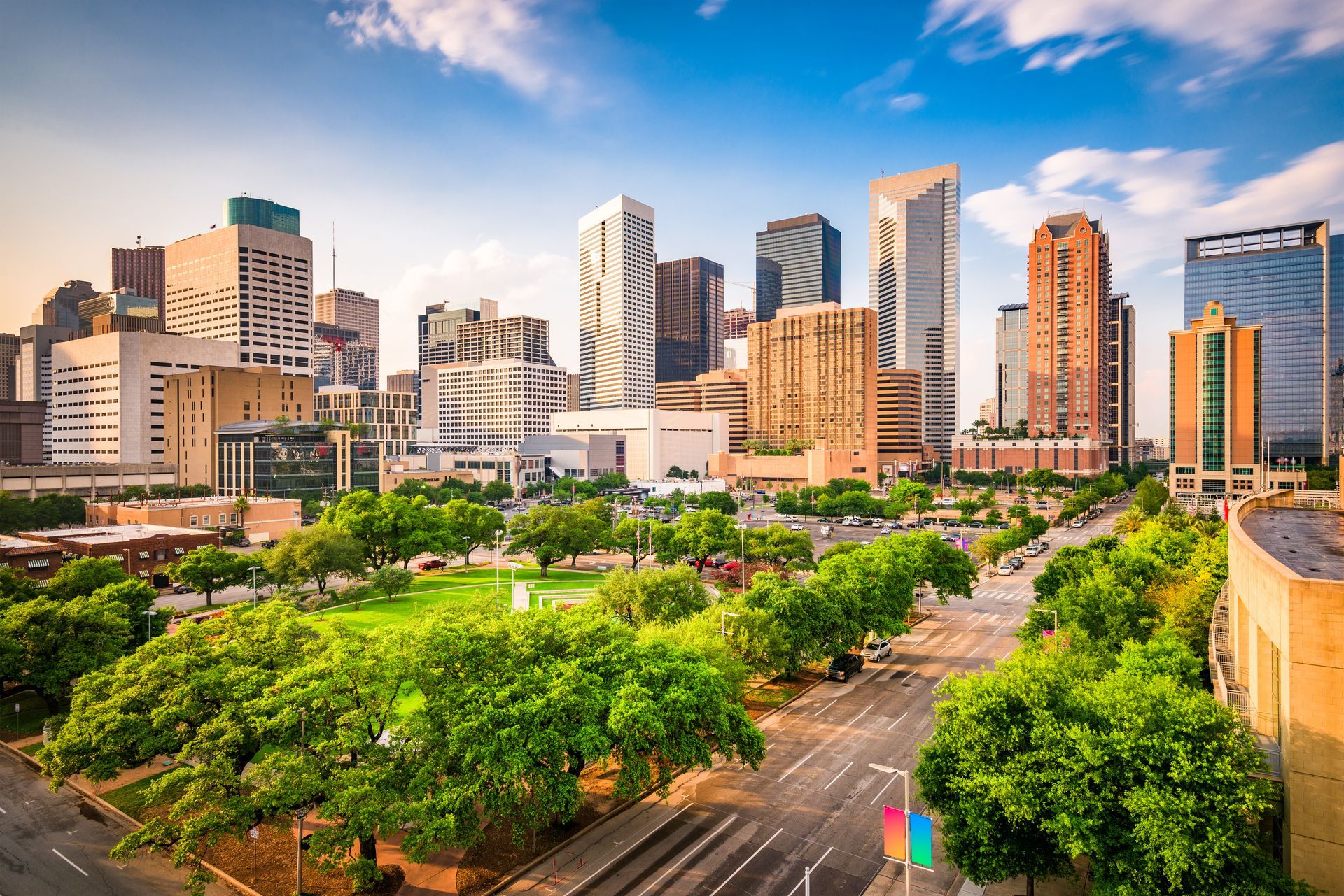 City skyline with modern skyscrapers above a green park and tree-lined road under a blue sky