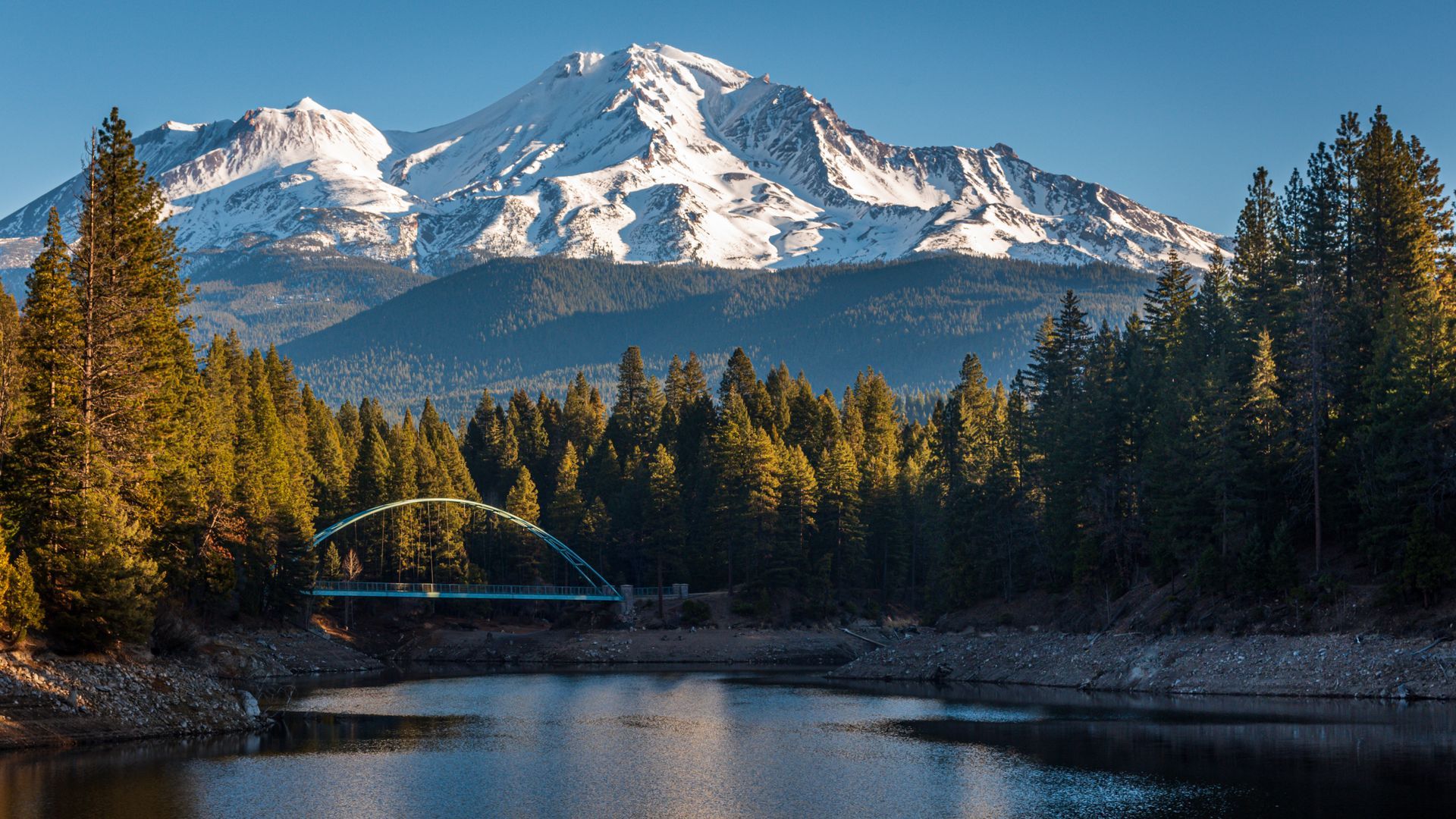 Snowy mountain above a forest and bridge over calm river at sunset