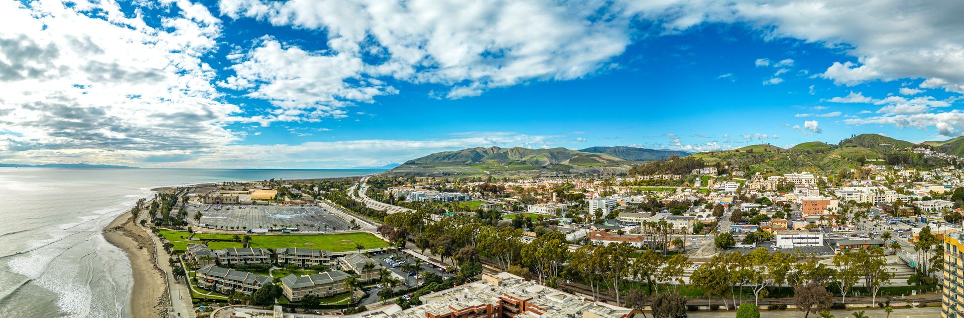 Panoramic coastal town with rocky shoreline, houses on hills, and a bright blue sky with clouds