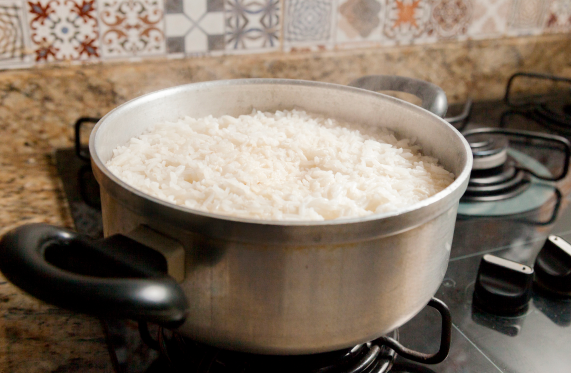 Panela de arroz branco cozinhando no fogão, com azulejos estampados na parede atrás da panela.