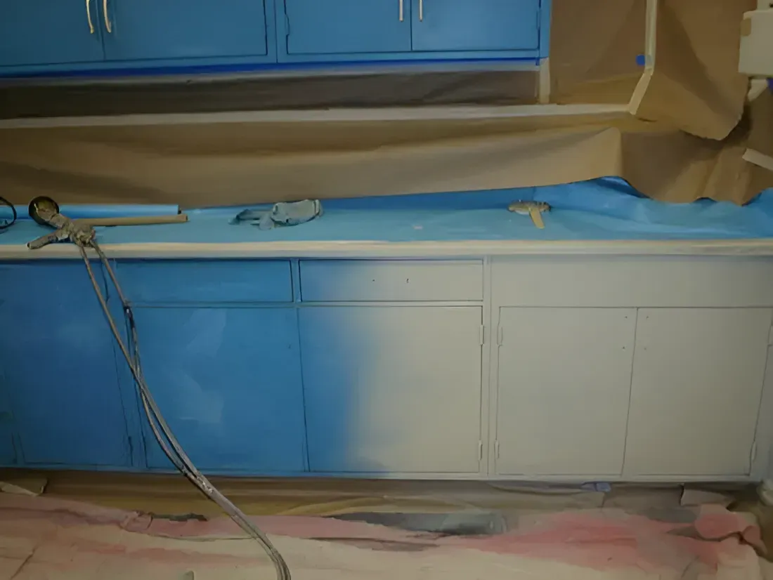 Blue and white kitchen cabinets being painted, with a paint sprayer and masking.
