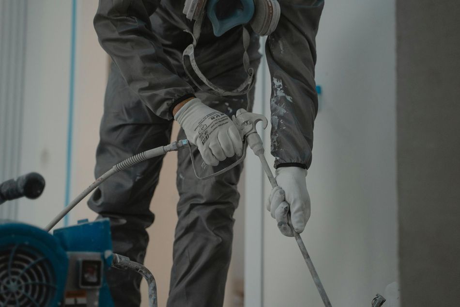 Person in protective suit and gloves using paint sprayer on a wall.