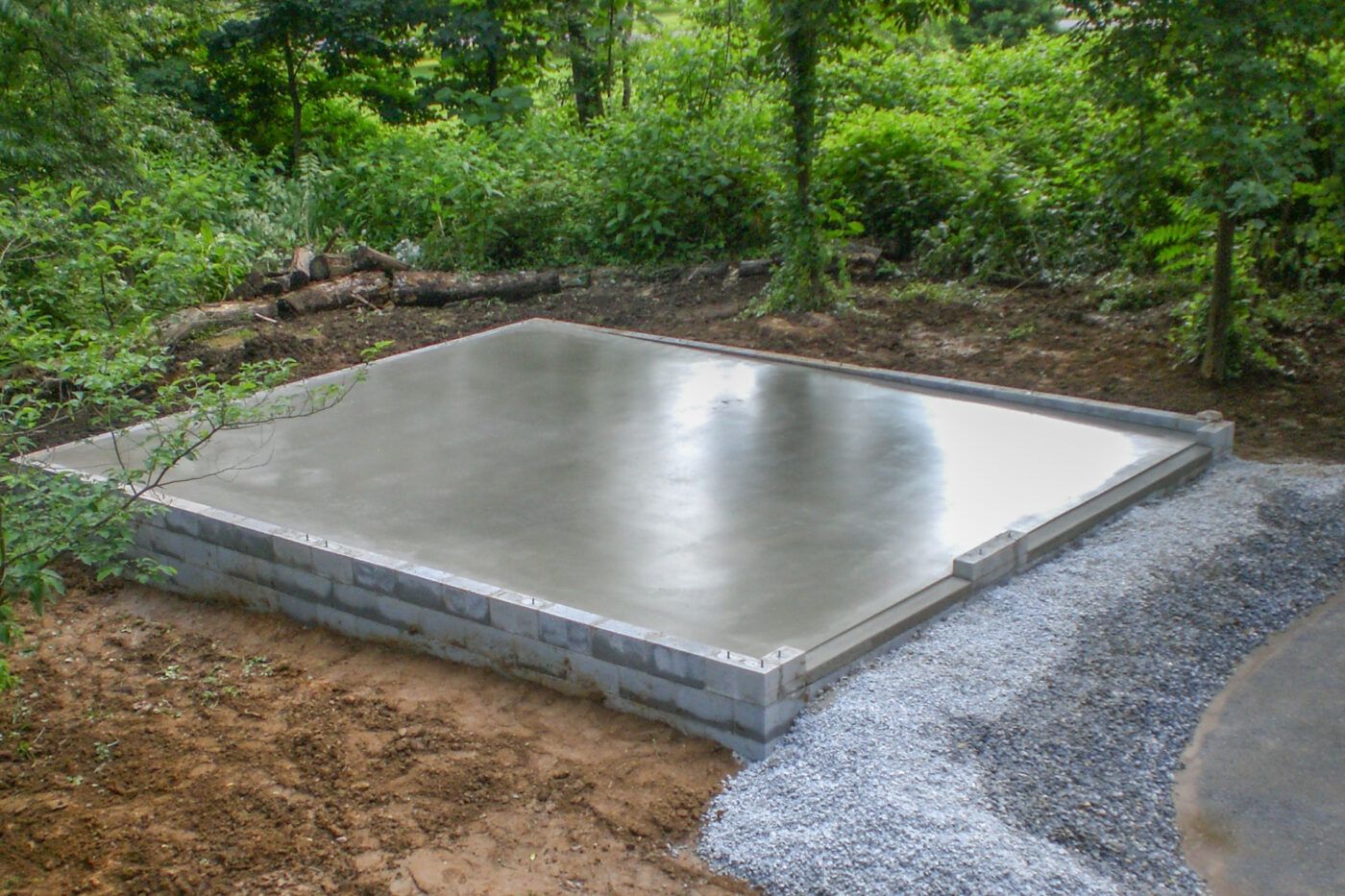 A newly poured concrete foundation slab elevated by concrete blocks, surrounded by gravel and a wooded landscape.
