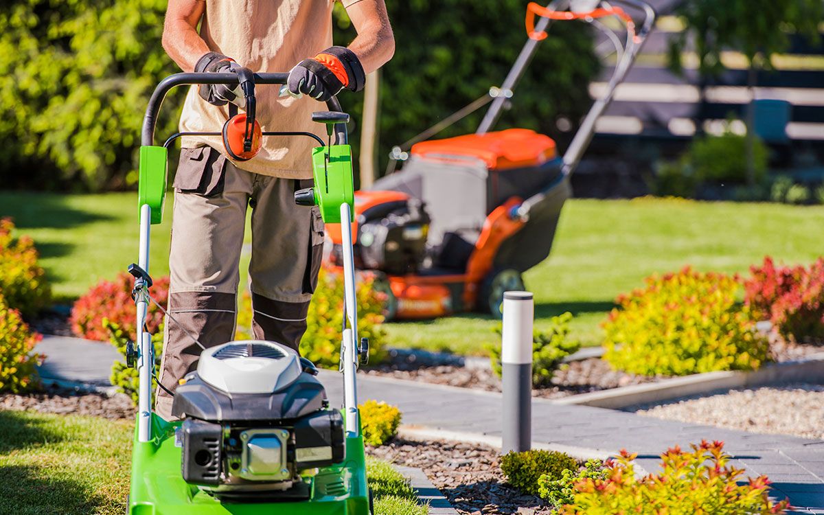 A person wearing work clothes and gloves pushes a green lawnmower in a landscaped garden with a second mower nearby.