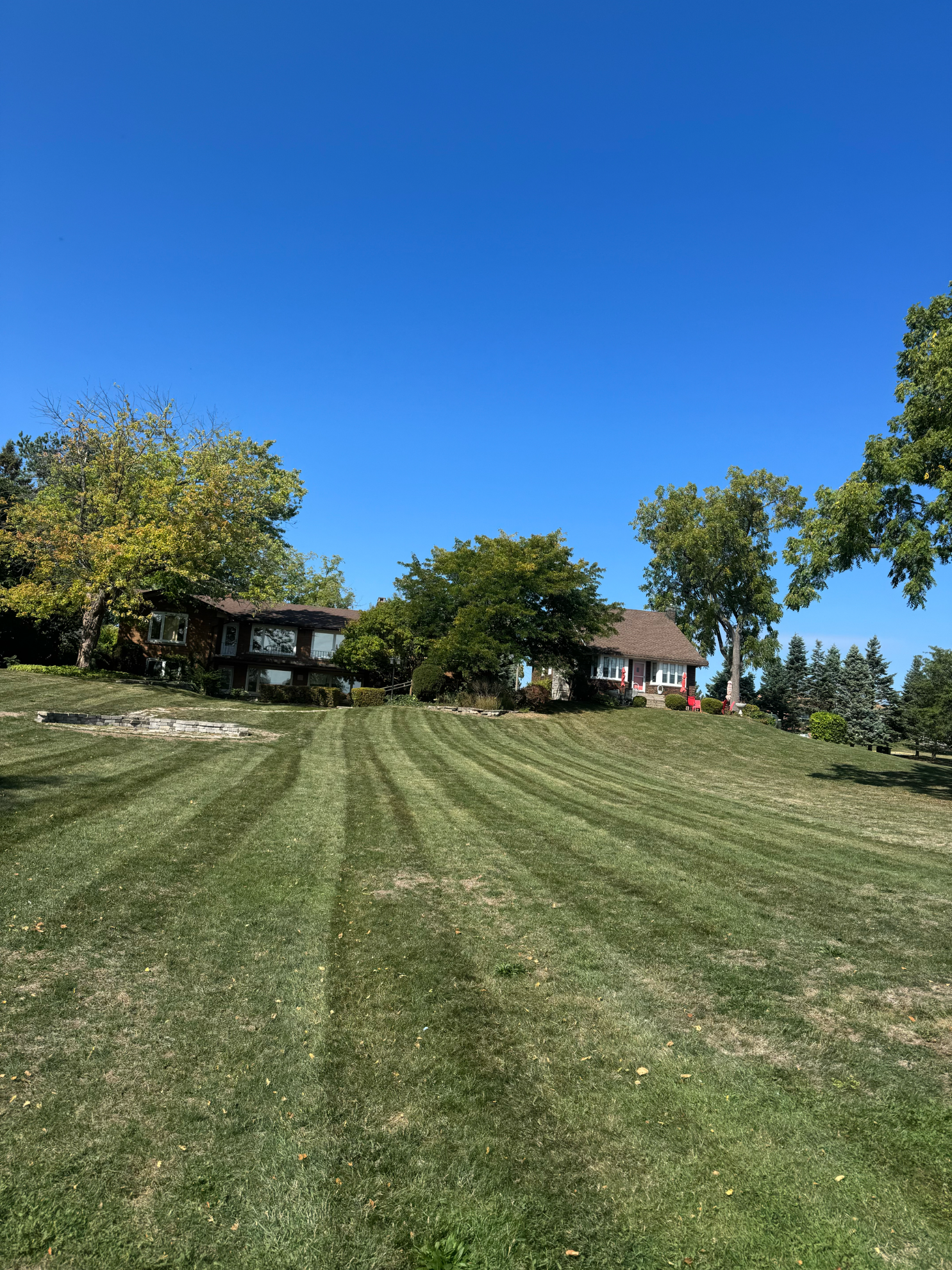 A house with a dark roof sits on a hill behind a large, neatly mowed lawn with prominent striped patterns under a blue sky.