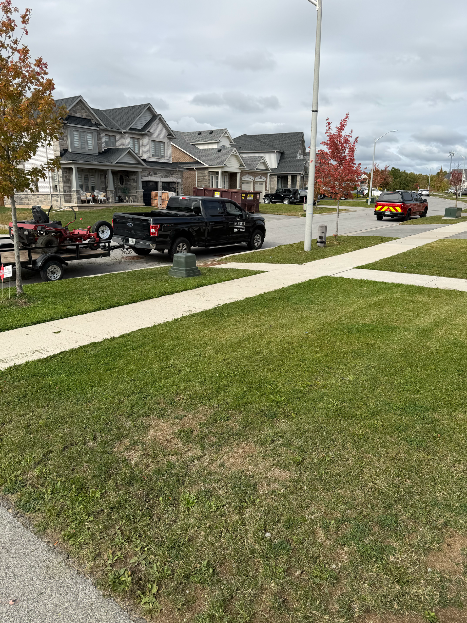 A black pickup truck towing a trailer with landscaping equipment parked on a residential street with houses in the background.