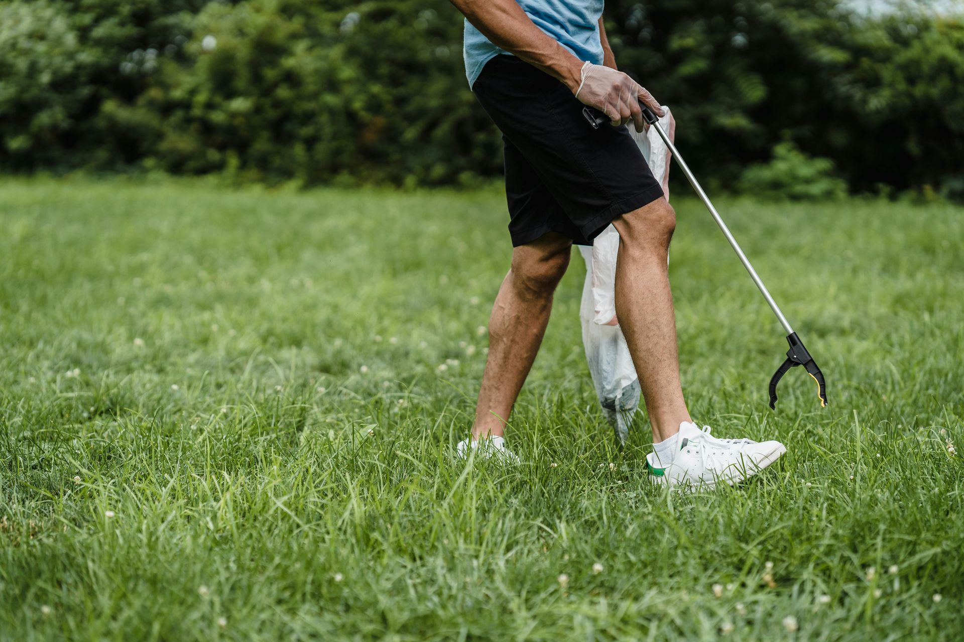 A person in a blue shirt and black shorts uses a reacher tool to collect litter in a grassy park.