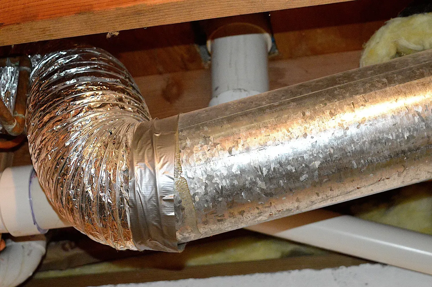 A silver flexible duct connected to a metal pipe under a wooden floor.