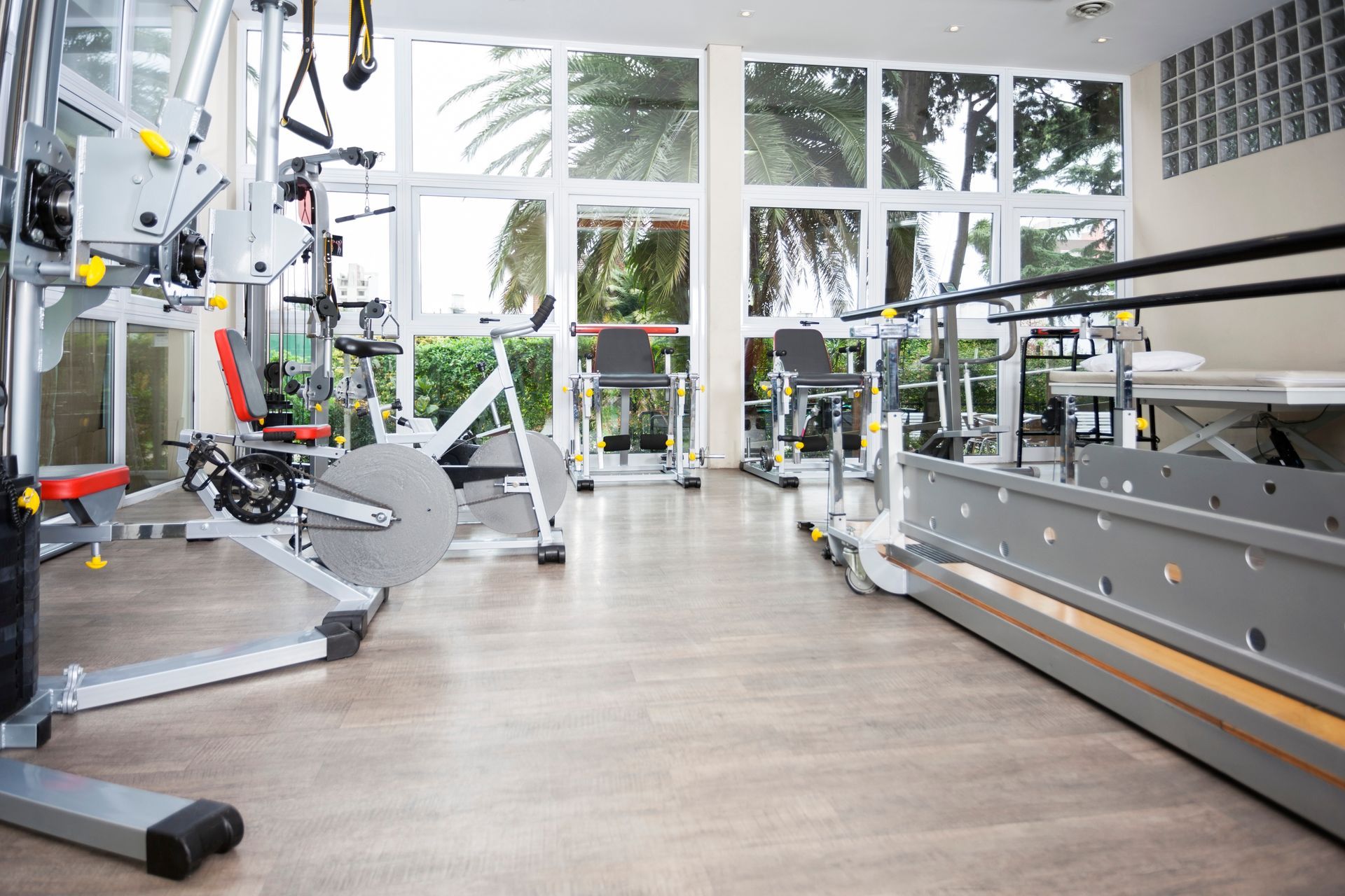A brightly lit fitness center with exercise machines and parallel bars, viewed from a low angle on a light wood floor.