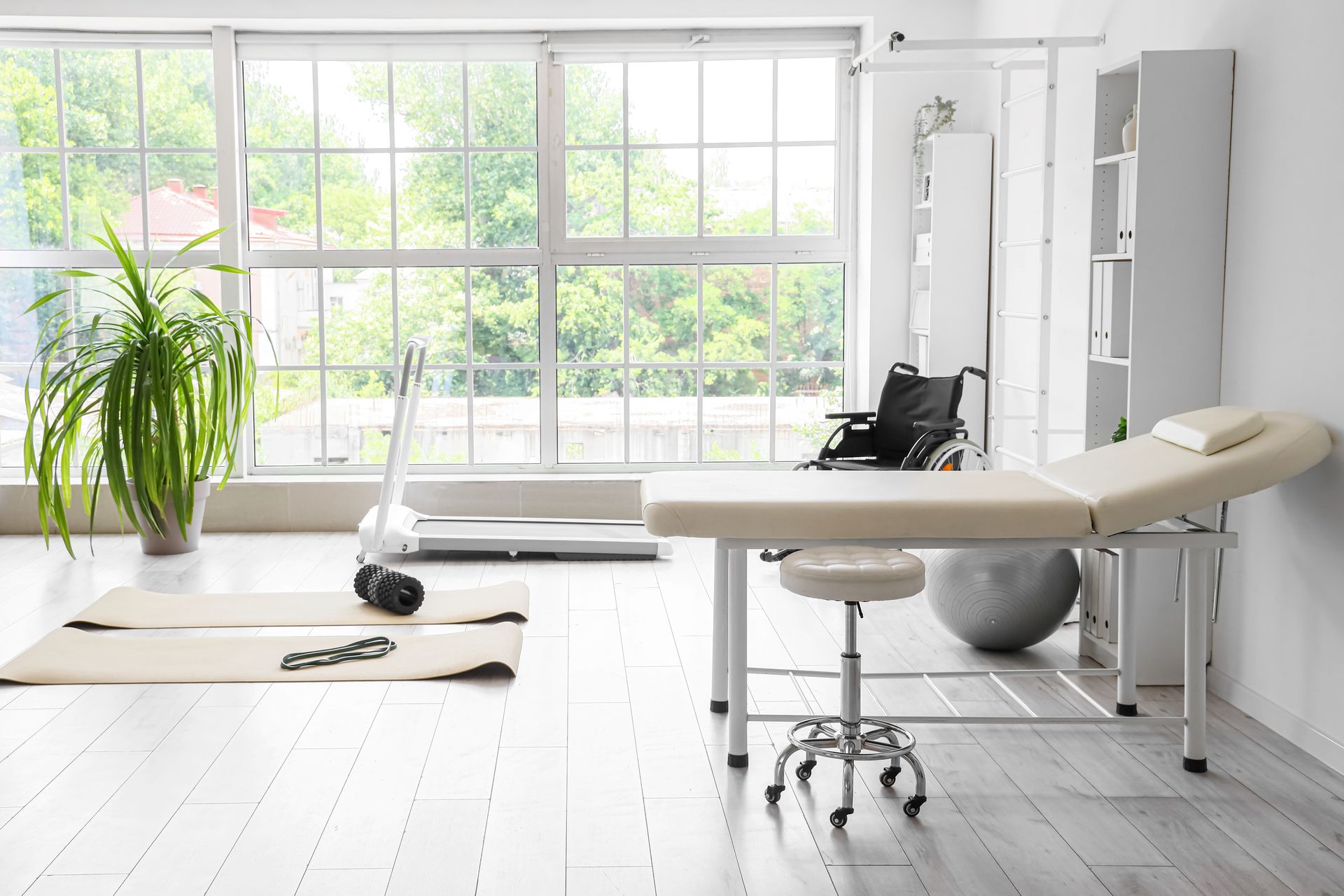 A bright physiotherapy or medical clinic room with a treatment table, exercise mats, a treadmill, and a wheelchair.
