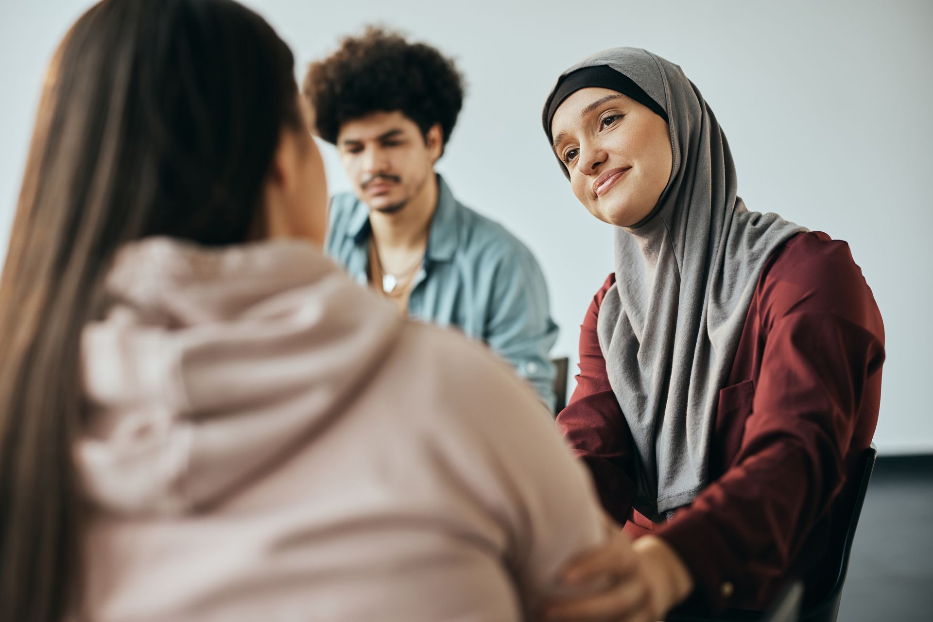 Three people seated in a group, with one person in a hijab smiling while touching the arm of the person in the foreground.
