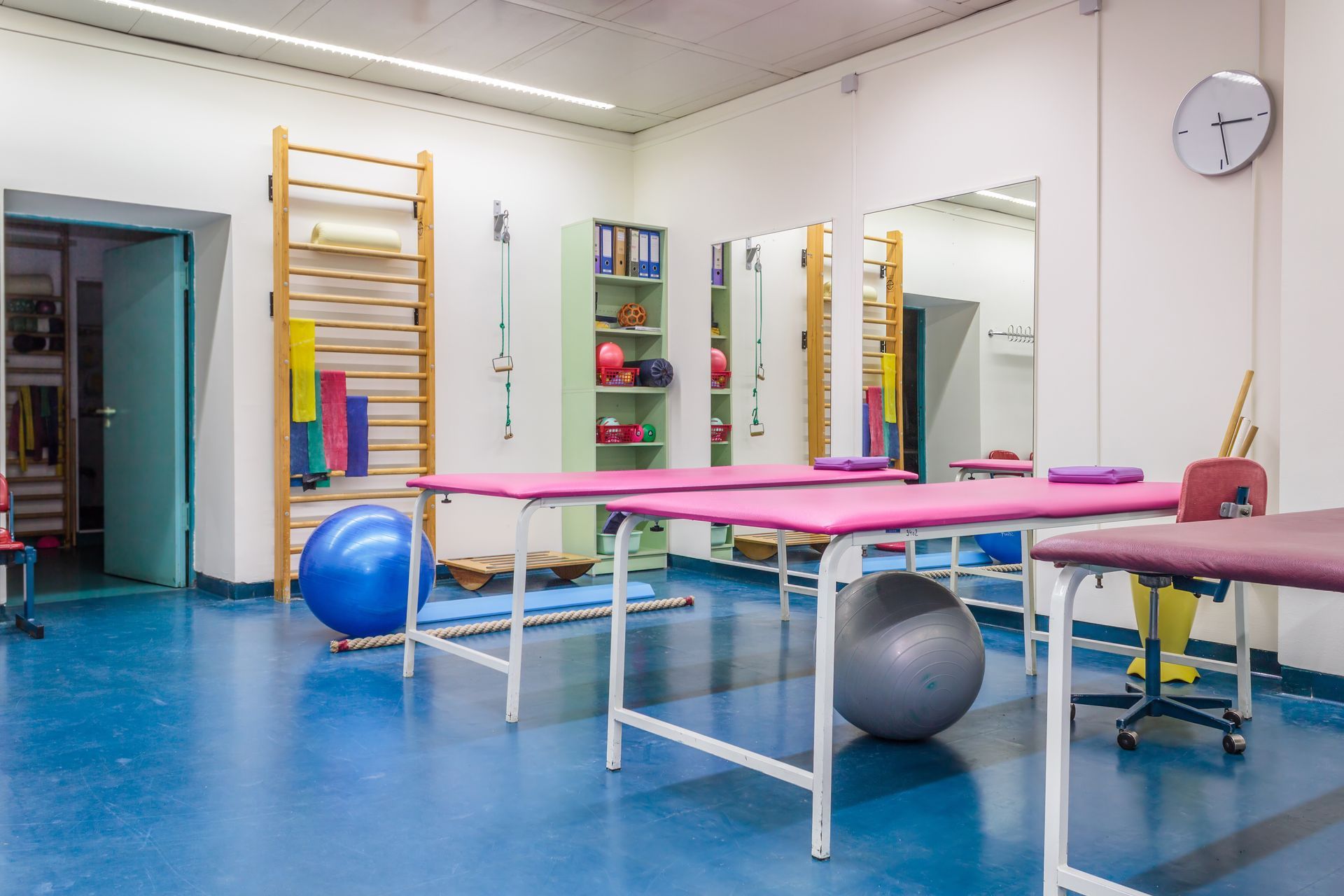 A physical therapy room with pink treatment tables, balance balls, and wall-mounted wooden exercise ladders. A physical therapy room with pink treatment tables, balance balls, and wall-mounted wooden exercise ladders.