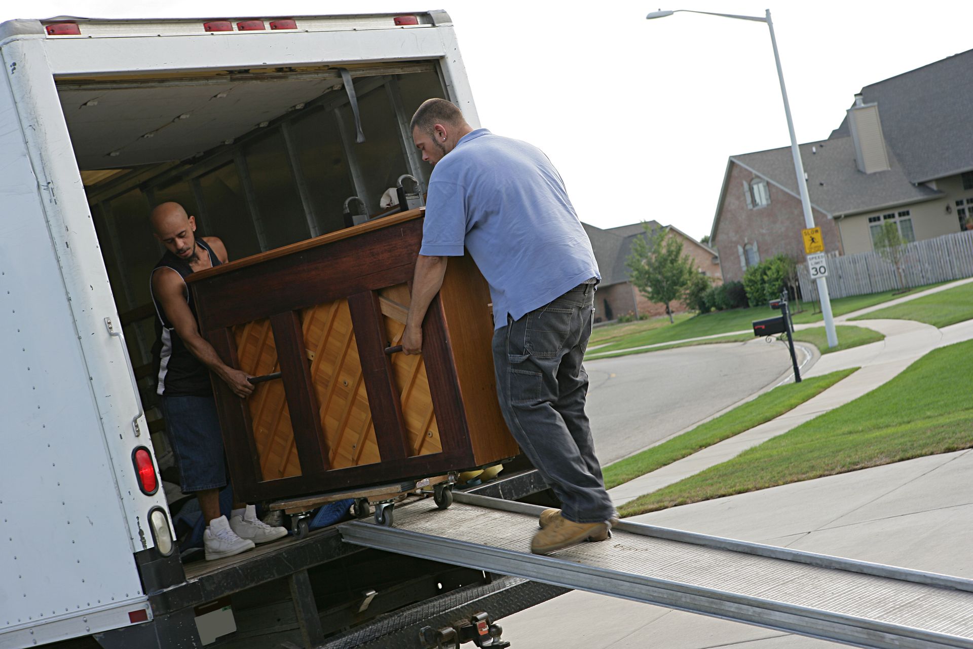 Two people loading a piano into a moving truck on a residential street.