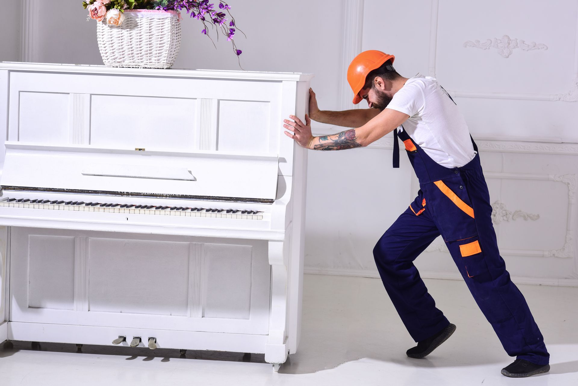 Man in orange hard hat pushing a white upright piano in a bright room