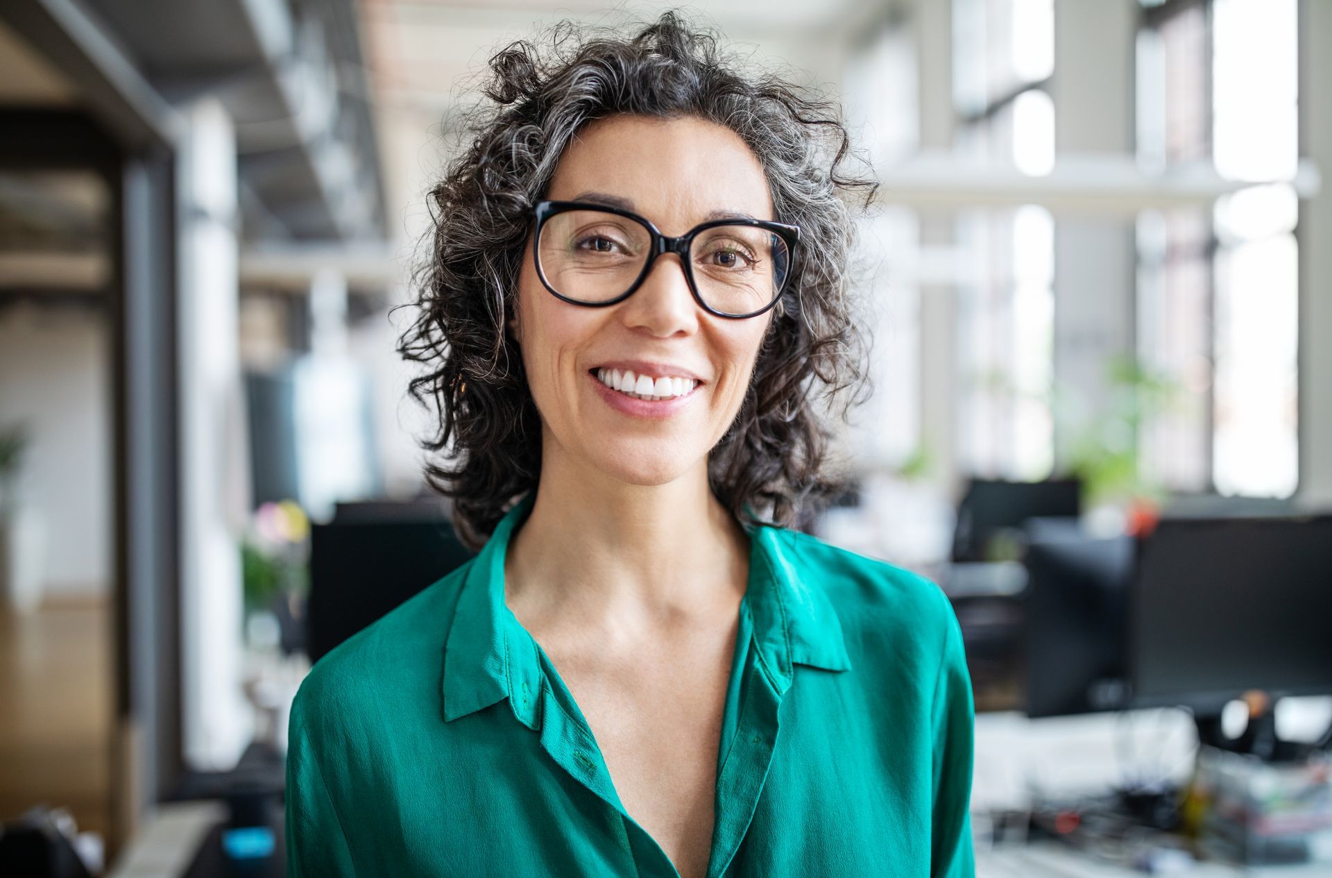 A woman wearing glasses and a green shirt is smiling in an office.