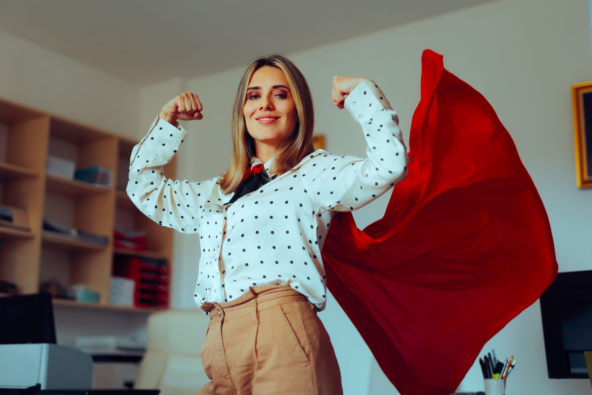 A woman is flexing her muscles in an office while wearing a red cape.