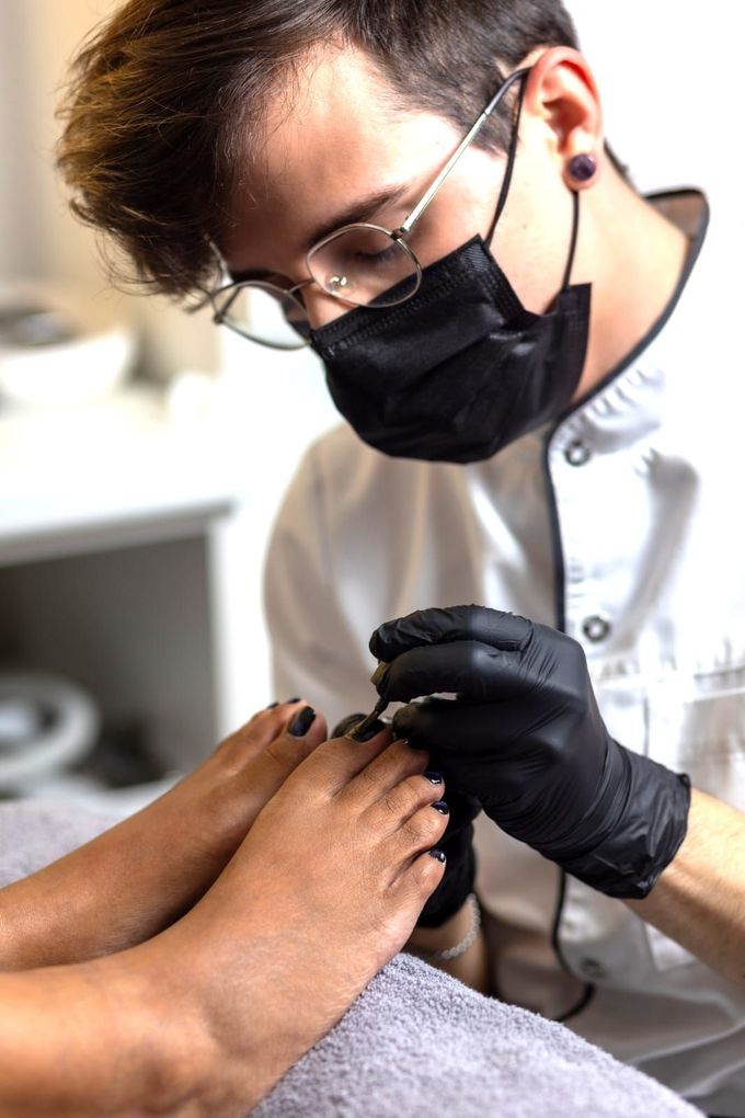 Ein Mann mit Maske und schwarzen Handschuhen lackiert einer Frau die Nägel.