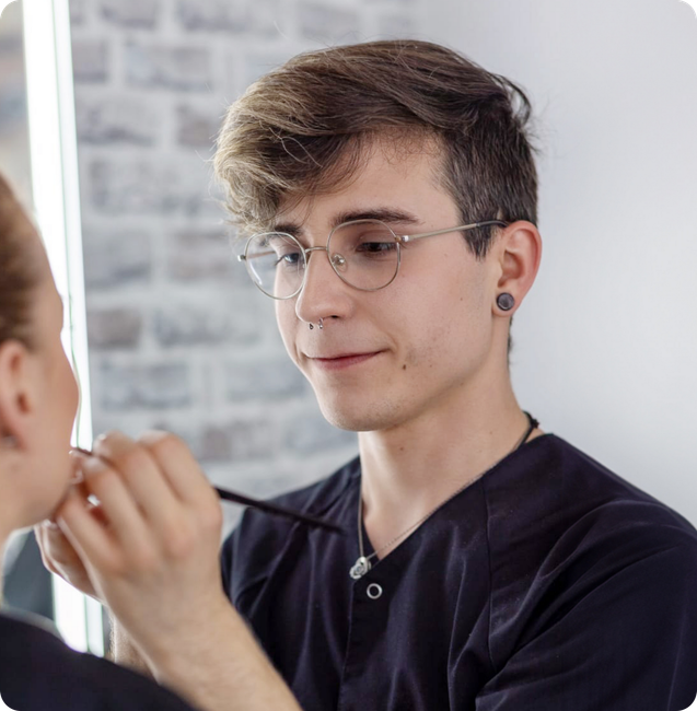 Ein Mann mit Brille schminkt das Gesicht einer Frau